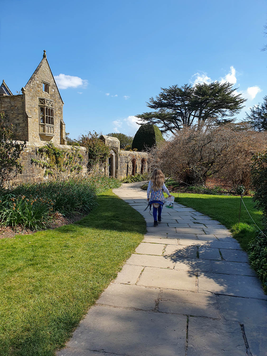 My daughter walks along a path towards the walled garden with part of the old house of Nymans seen in the background - visiting Nymans with kids