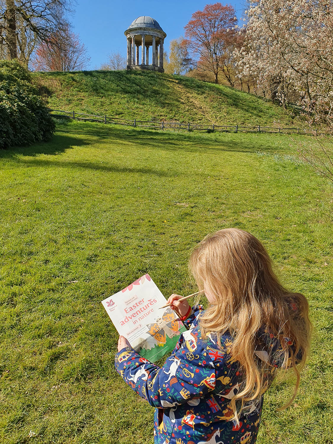 My daughter looks at her Easter adventure trail in the gardens of Petworth House with a Doric temple in the background - visiting Petworth with kids