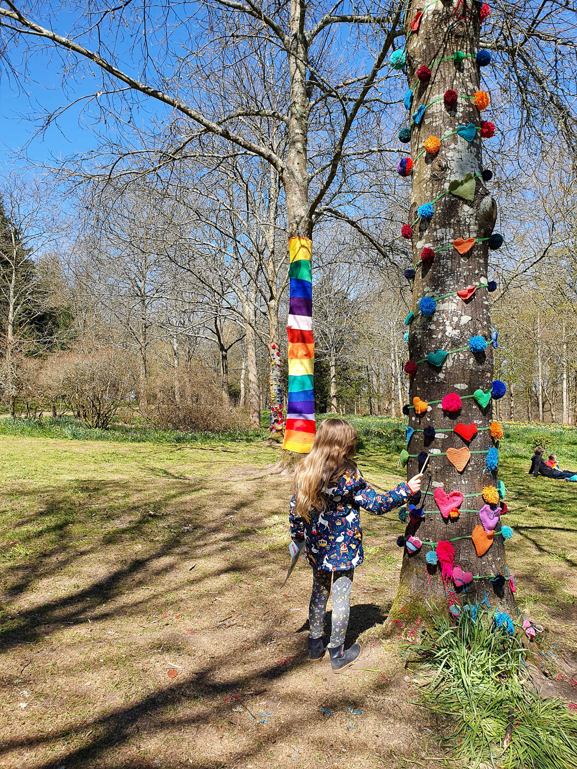 My daughter reaches her hand out to a tree covered in pompoms and shapes with another tree covered in bright stripes of wool as part of a yarnbombing project at Petworth - exploring the gardens and park at Petworth with kids
