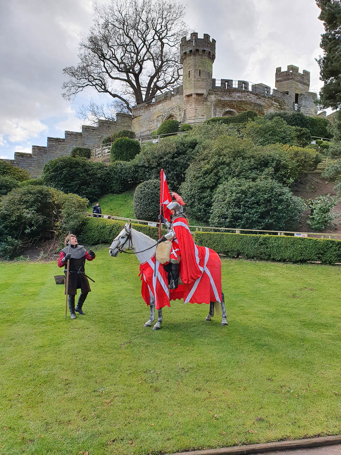 Actor playing the Earl of Warwick in armour on a white horse with another actor next to him and the Conqueror's Castle in the background - a day out at Warwick Castle with kids, including the new Zog trail