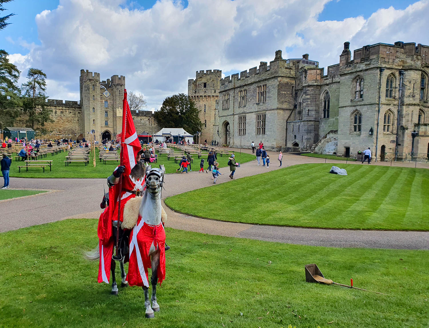 Armoured actor playing the Earl of Warwick on his white horse with the inner court of Warwick Castle in the background, including picnic benches on the grass