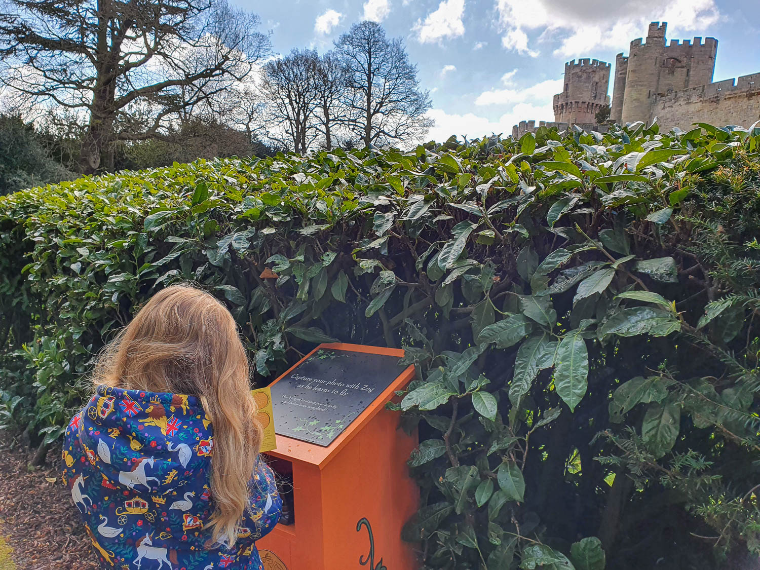 My daughter looks at her trail card by the stamp machine on the Zog trail at Warwick Castle, with the castle towers behind a hedge in the background - my Warwick Castle Zog trail review