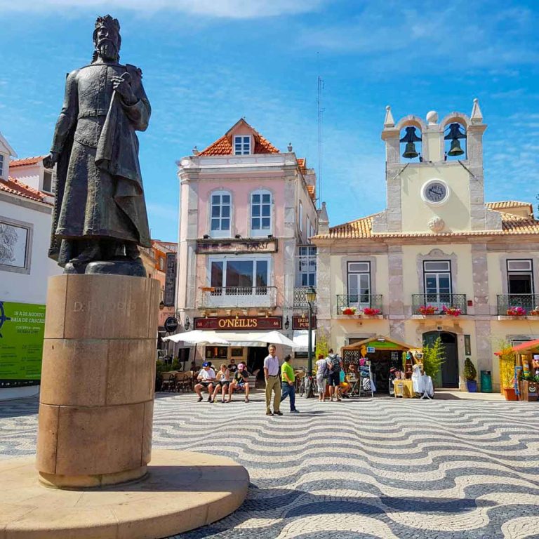 A square in Cascais with black and white wave pattern tiles, statue and brightly coloured buildings under a blue sky - remembering some of my favourite memories of the country and Portugal's most unusual attractions