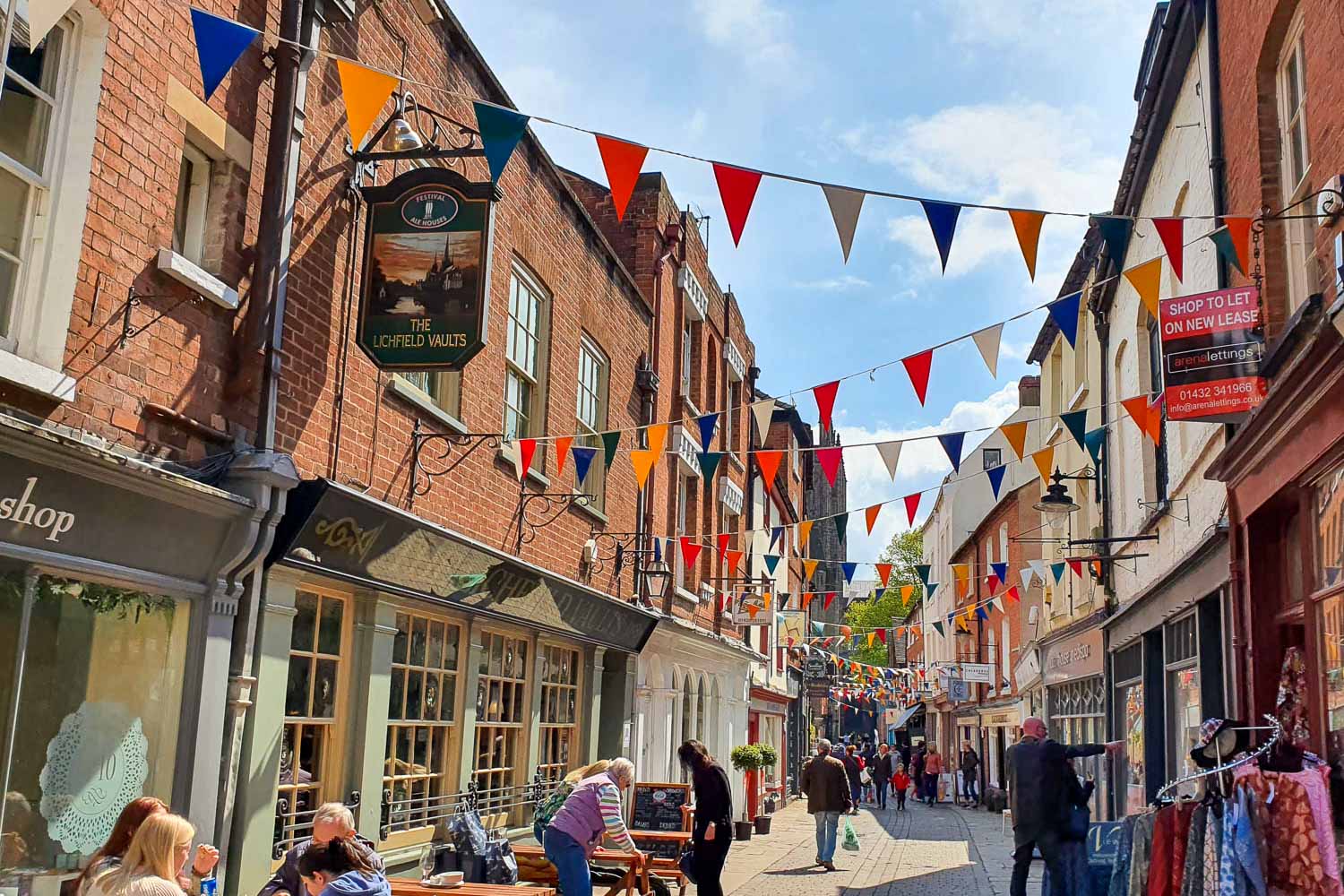View along Church Street in Herefords with bunting strung between the shops above the tables of cafes on a sunny day - a great place to pick up food during a day in Hereford with kids