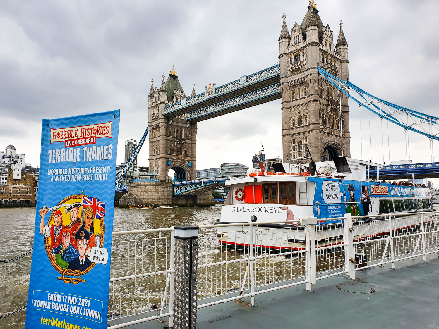 View of a sign for the Terrible Thames boat tour and the boat itself, with Tower Bridge in the background on a cloudy grey day - one of the best boat tours in London with kids