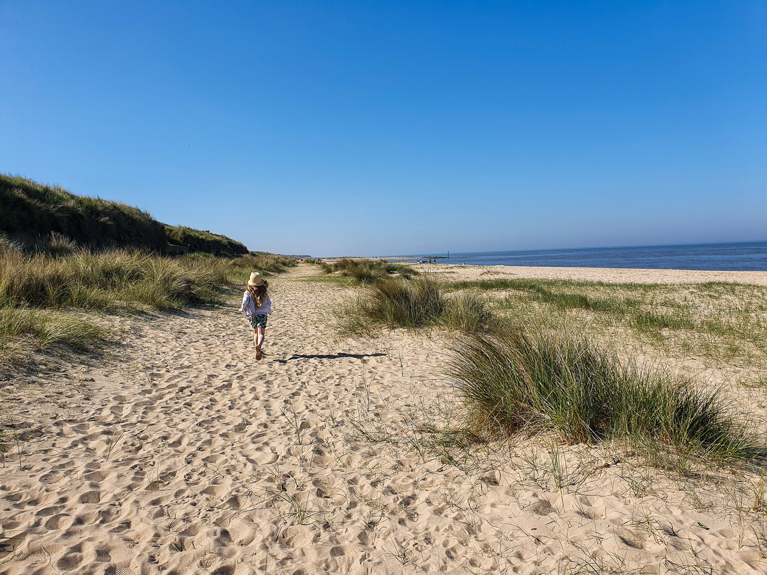 My daughter walks along the sand by the dunes at Caister on Sea beach, one of the best beaches in Norfolk with kids if you're looking to escape the crowds