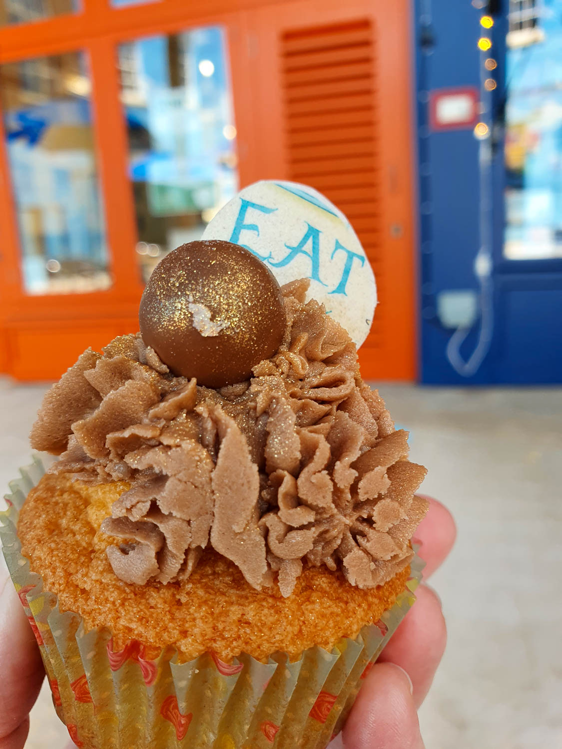 Hang holding a cupcake with chocolate icing and gilded malteser with a sign saying eat me plus the orange and blue front of the cafe at the Oxford Story Museum in the background - my review of a family day out in Oxford
