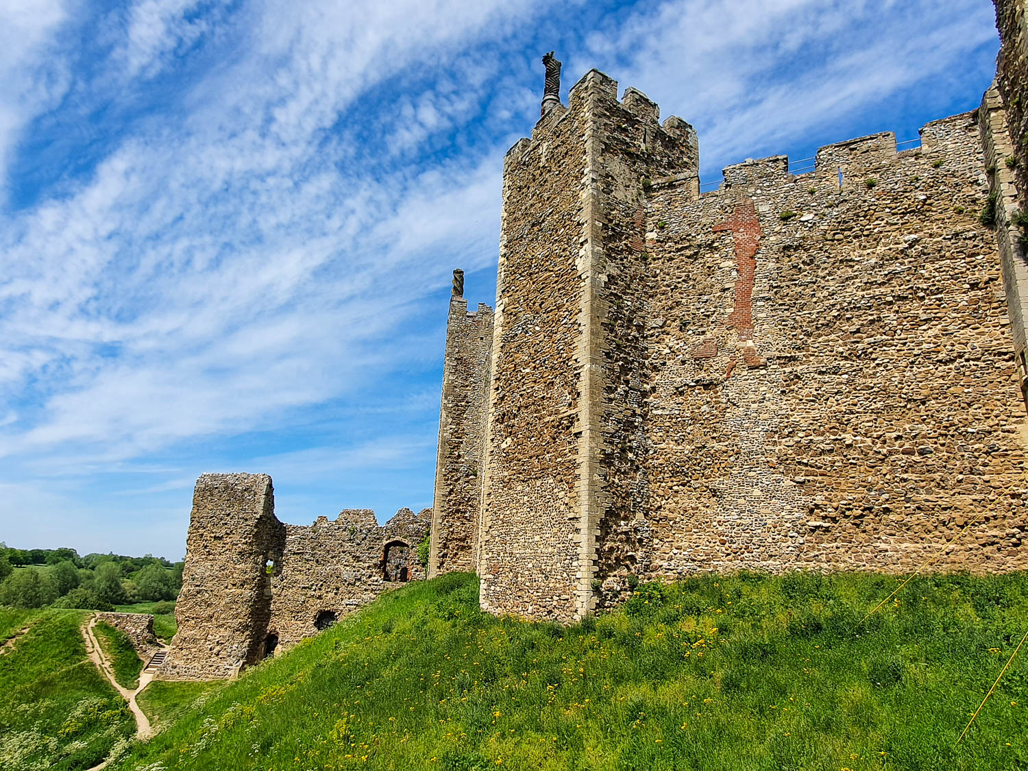 View of the towers and exterior of Framlingham Castle against a blue sky, taken from the moat, with the walking paths leading off behind the walls - open to the public to explore if you're visiting Suffolk with kids