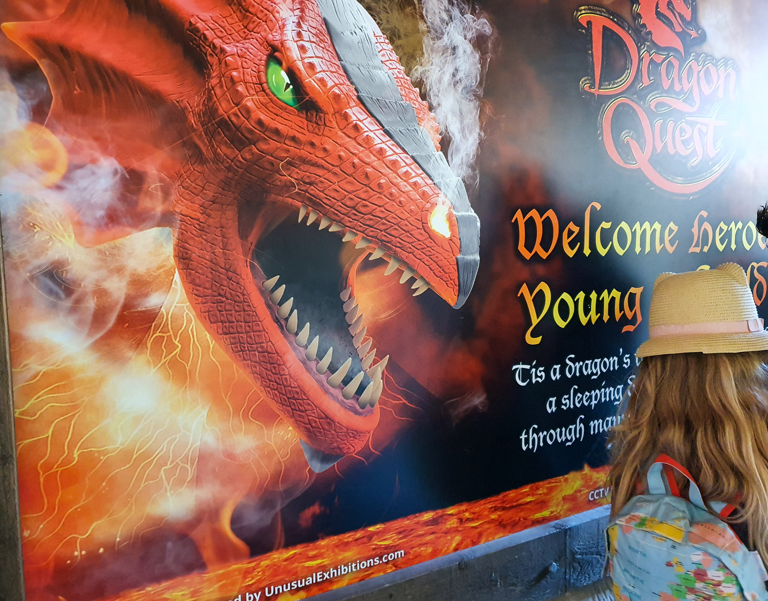 The sign for Dragon Quest at Alnwick Castle with a fire breathing scaly red dragon and a girl looking at the words - my review of visiting Alnwick Castle with kids