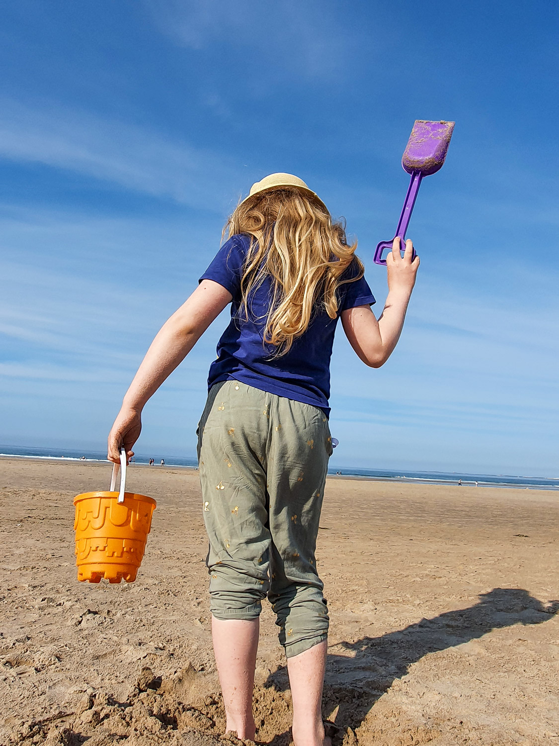 My daughter stands on Bamburgh beach with an orange bucket and purple spade, the sea visible on the horizon after a visit to Bamburgh Castle with kids