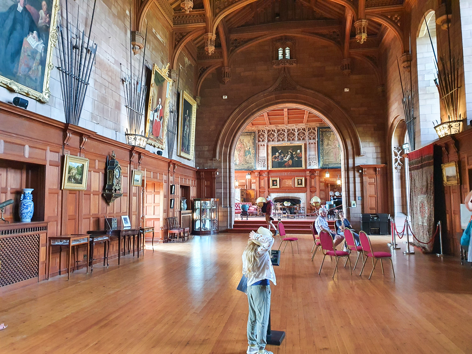 My daughter looks along the length of the King's Hall at Bamburgh Castle, with weapons and artwork on the walls and a high carved ceiling - my review of our day at Bamburgh Castle with kids