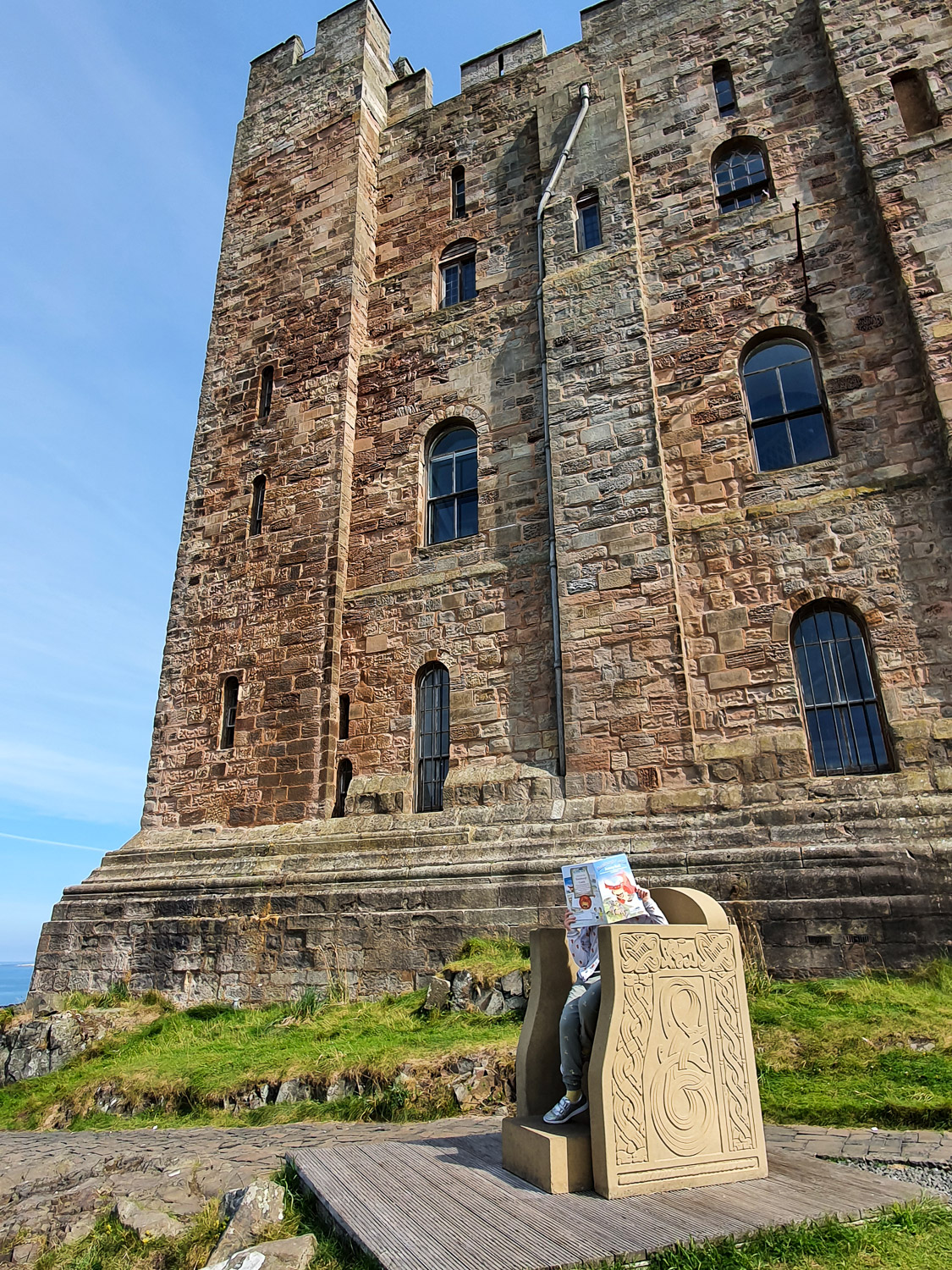 My daughter sits in the replica carved stone throne in the shadow of the huge square Norman keep - visiting Bamburgh Castle with kids
