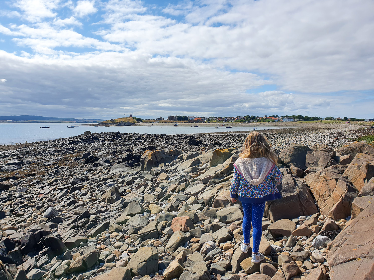 My daughter walks across the rocks on the seafront near the castle looking towards the harbour during a day on Lindisfarne with kids