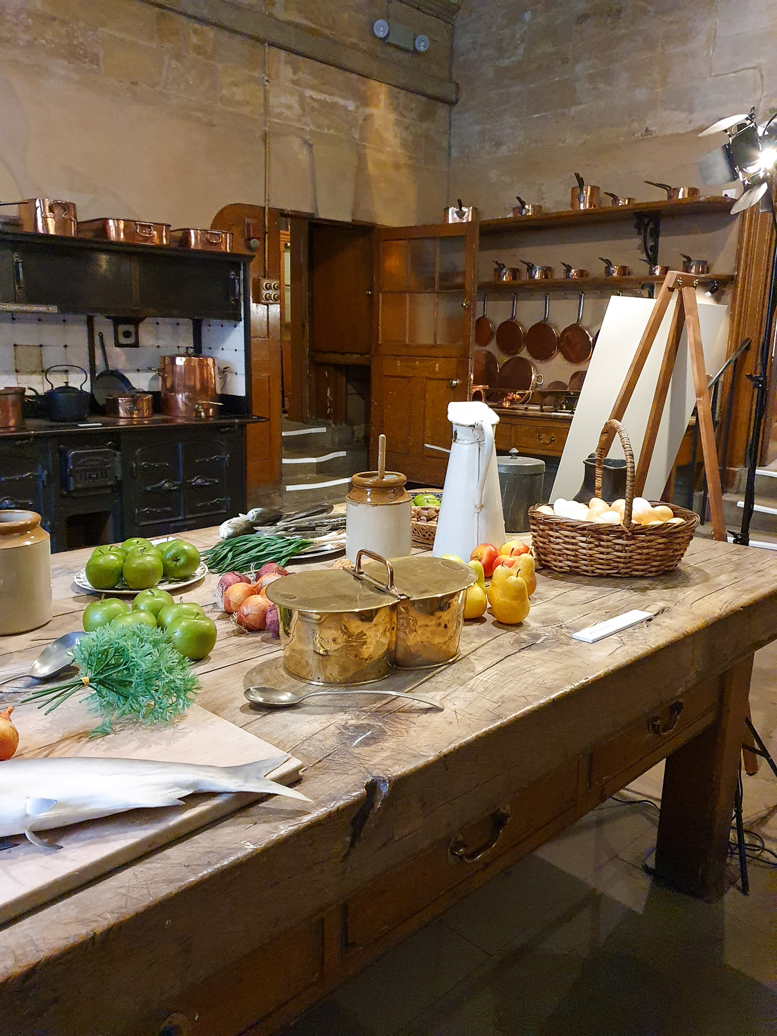 Old wooden table with convincing replica food including a fish and apples in the kitchens of Harewood House, with copper pans seen hanging in the background - my review of a day out at Harewood House with kids