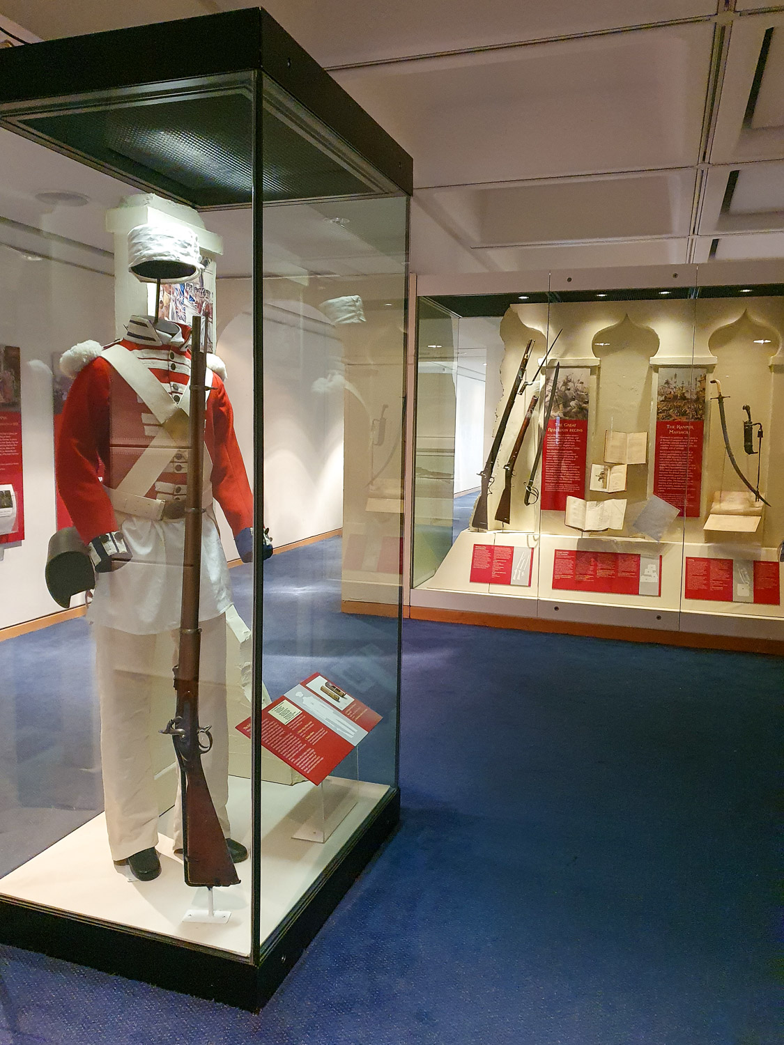 Glass display cases from the temporary exhibition on the Indian Mutiny, also known as the Great Rebellion, with a red and white British Empire uniform and guns and sword seen in the background - my review of a day out at the Royal Armouries with kids