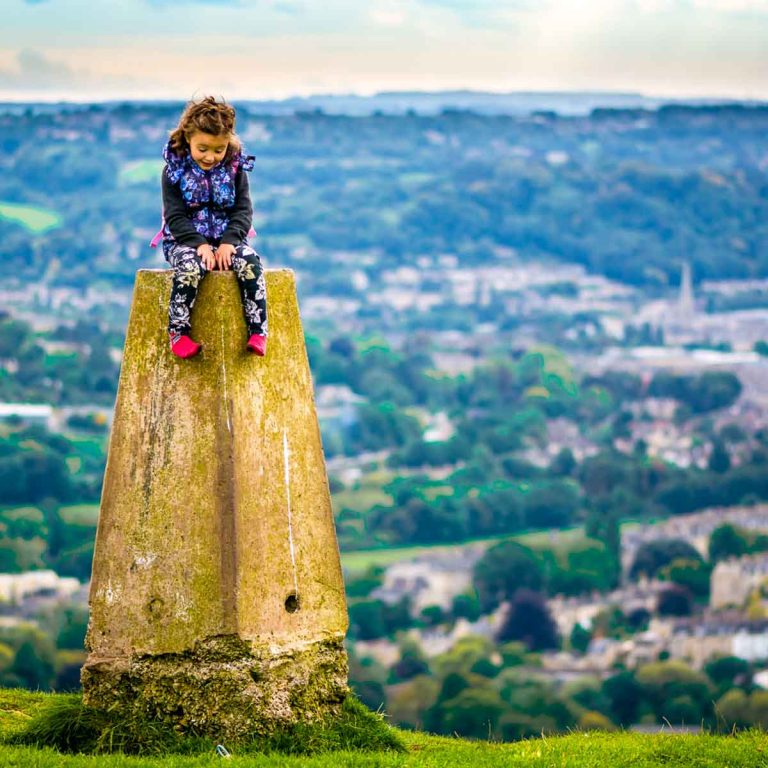 Girl sits on the top of a trig point, or triangulation pillar, on Solsbury Hill with Bath and the Somerset countryside in the background - discovering the best things to do in Somerset with kids