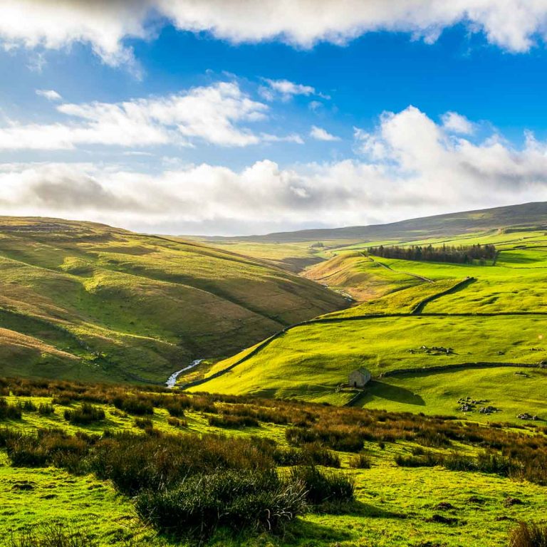 View across the Yorkshire Dales on a sunny day with a river and stone building just seen among the hills - the best things to do in Yorkshire with kids