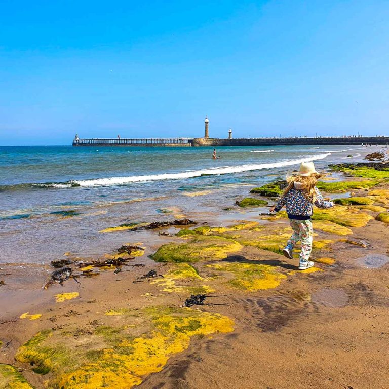 My daughter walks along West Cliff beach, with the lighthouses on the pier visible in the distance - my pick of the best things to do in Whitby with kids