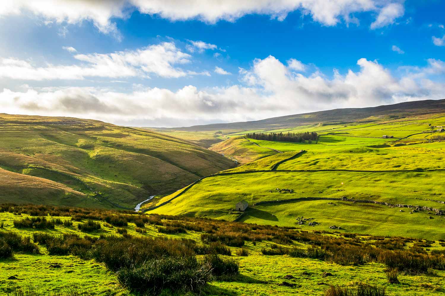View across the Yorkshire Dales on a sunny day with a river and stone building just seen among the hills - the best things to do in Yorkshire with kids