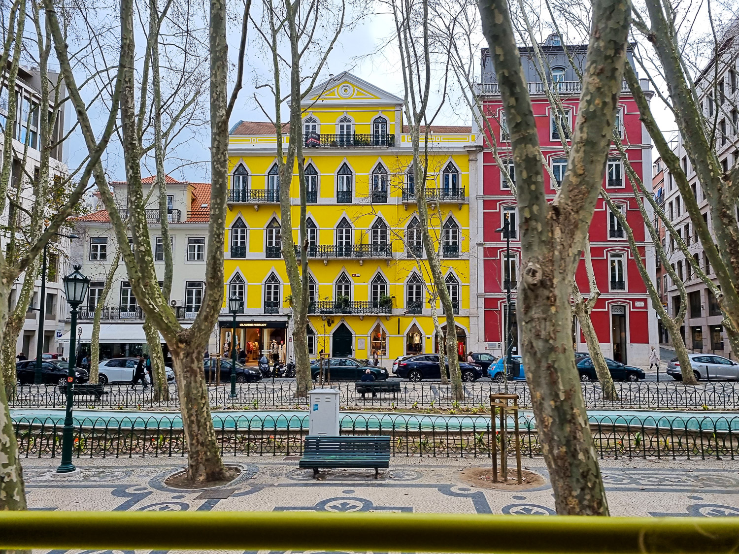 View of bright yellow and red buildings seen through the trees on a square in Lisbon, viewed over the yellow bar at the side of our Hippotrip amphibious vehicle during a tour of Lisbon with kids