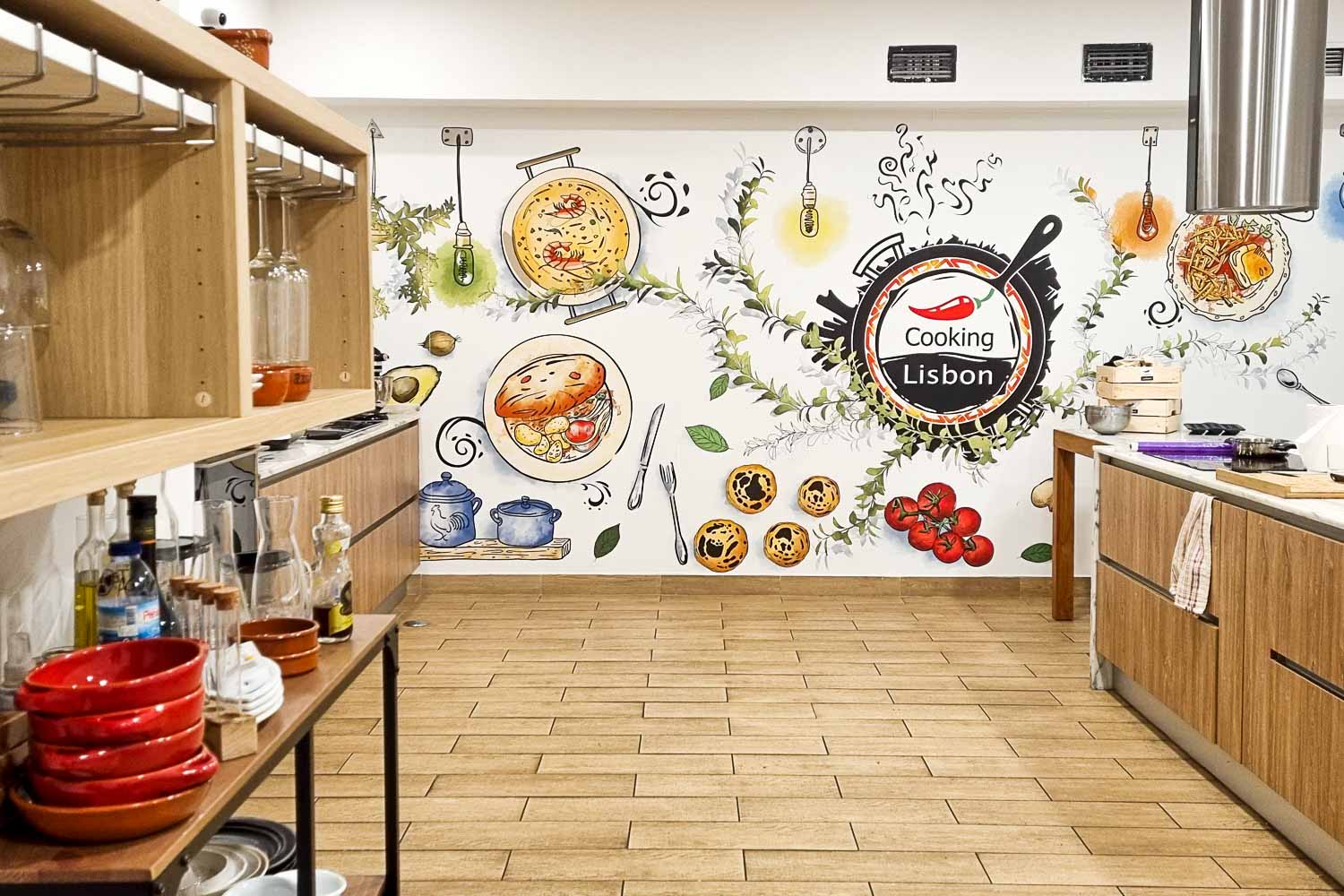 View of the Cooking Lisbon kitchen with bowls, oil and other ingredients on the shelves, bright mural on the wall, and worktops just seen - my review of our Lisbon cooking class with kids