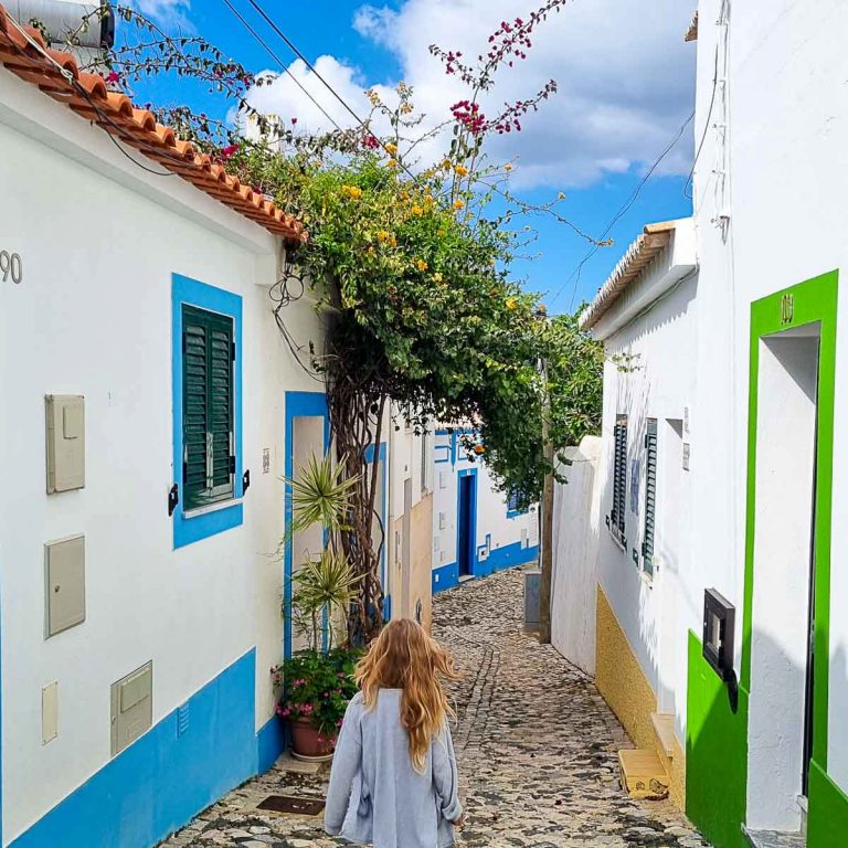 My daughter walks down a street in Ferragudo with white houses decorated with light blue and bright green and a flowering tree above the street - the best things to do in the Algarve with kids