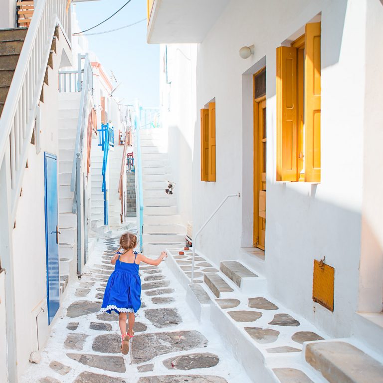 Girl in blue dress runs along paved street between white houses in Mykonos - my guide to Greece with kids
