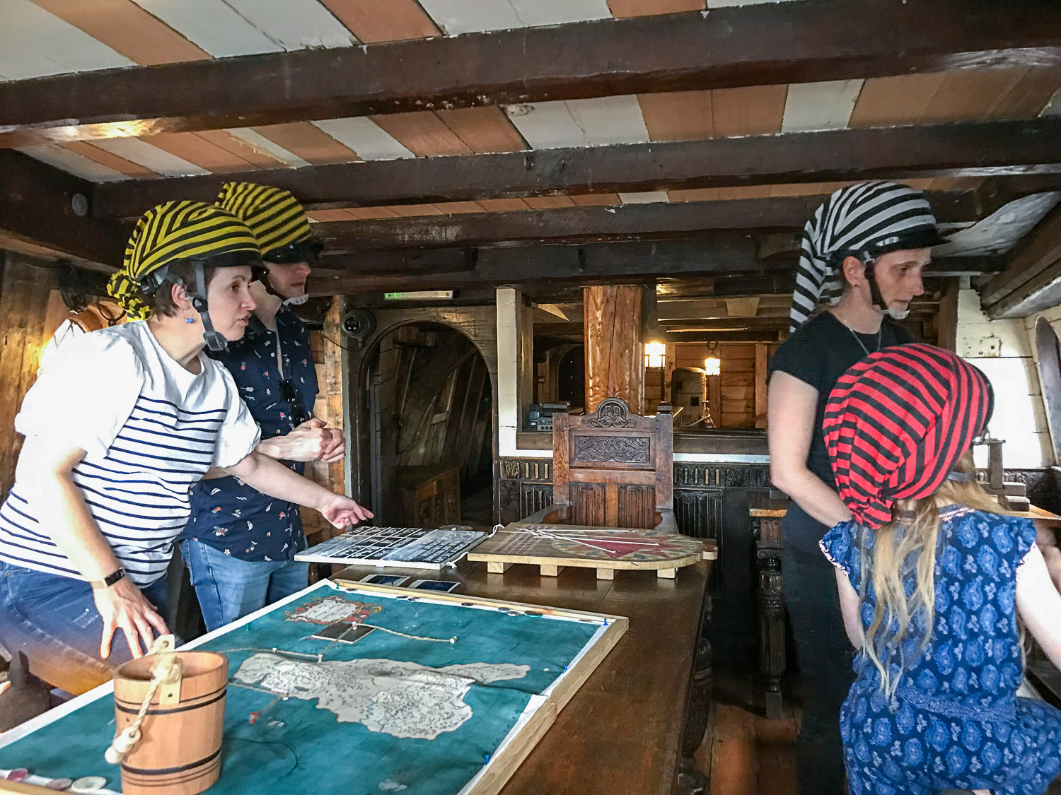 Three adults and a child all trying to solve a puzzle, with various maps and other items laid out on the table of the ship's cabin during the Golden Hinde escape room with kids - my Escape from the Golden Hinde review