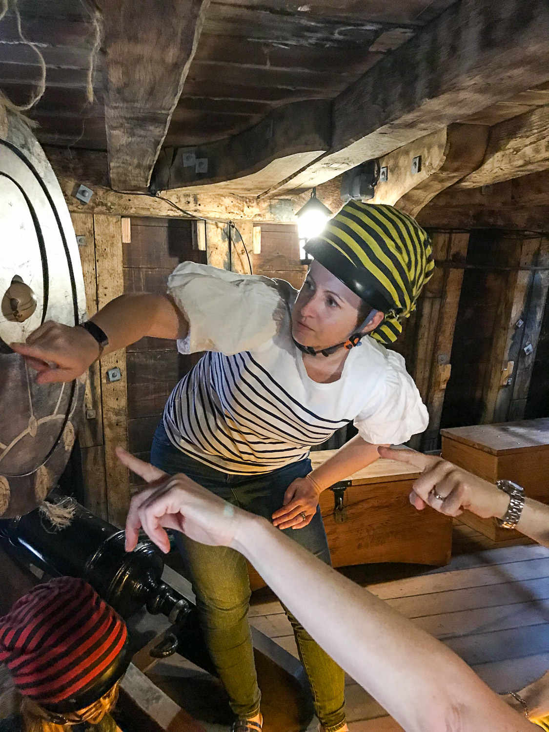 Woman bending down to point at a hanging circle of wooden with other hands also pointing the same direction, during the Golden Hinde escape room game - my review of Escape from the Golden Hinde with kids