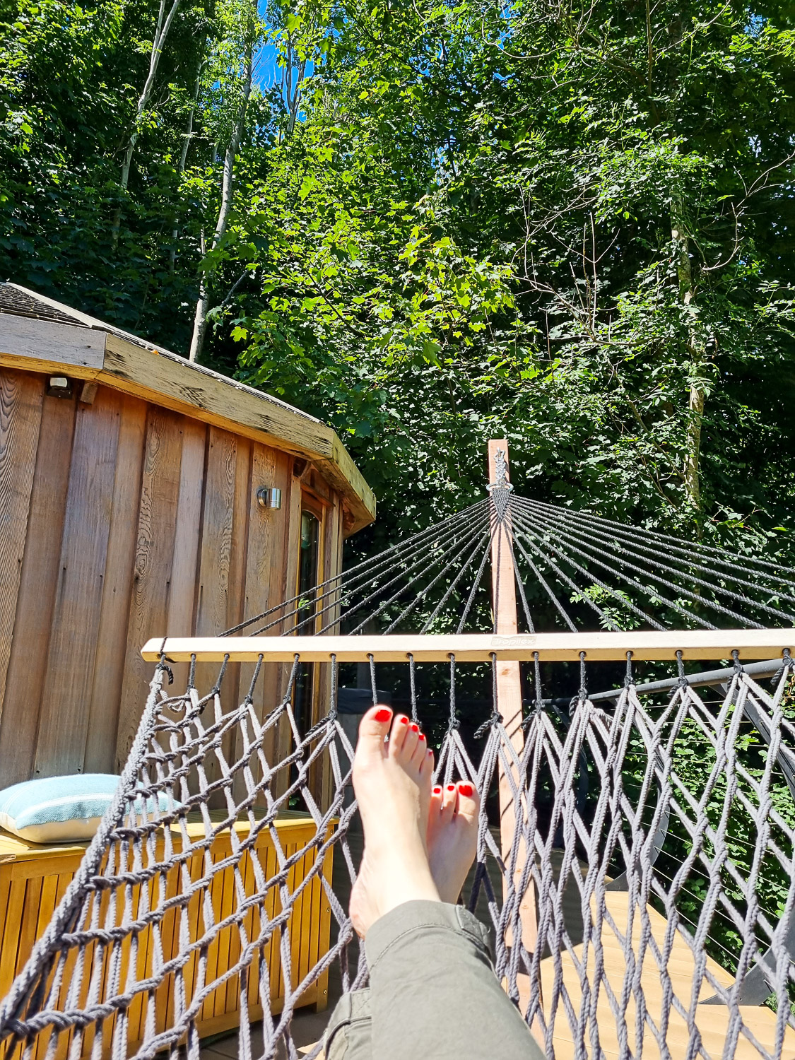 Woman's legs in khaki trousers and bare feet in the end of a rope hammock with wooden building seen among the trees - my Dittisham Hideaway review during a Devon luxury glamping break