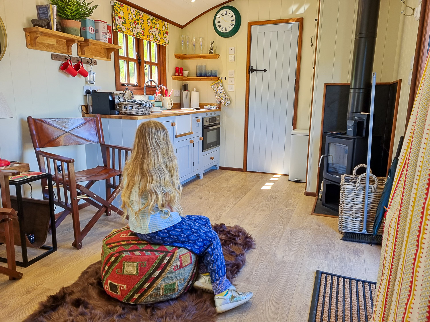 My daughter sits on the pouffe in the living space looking to the kitchen, with stove and bathroom door beyond, in one of the shepherd's huts at Dittisham Hideaway - my review of the Devon luxury glamping site
