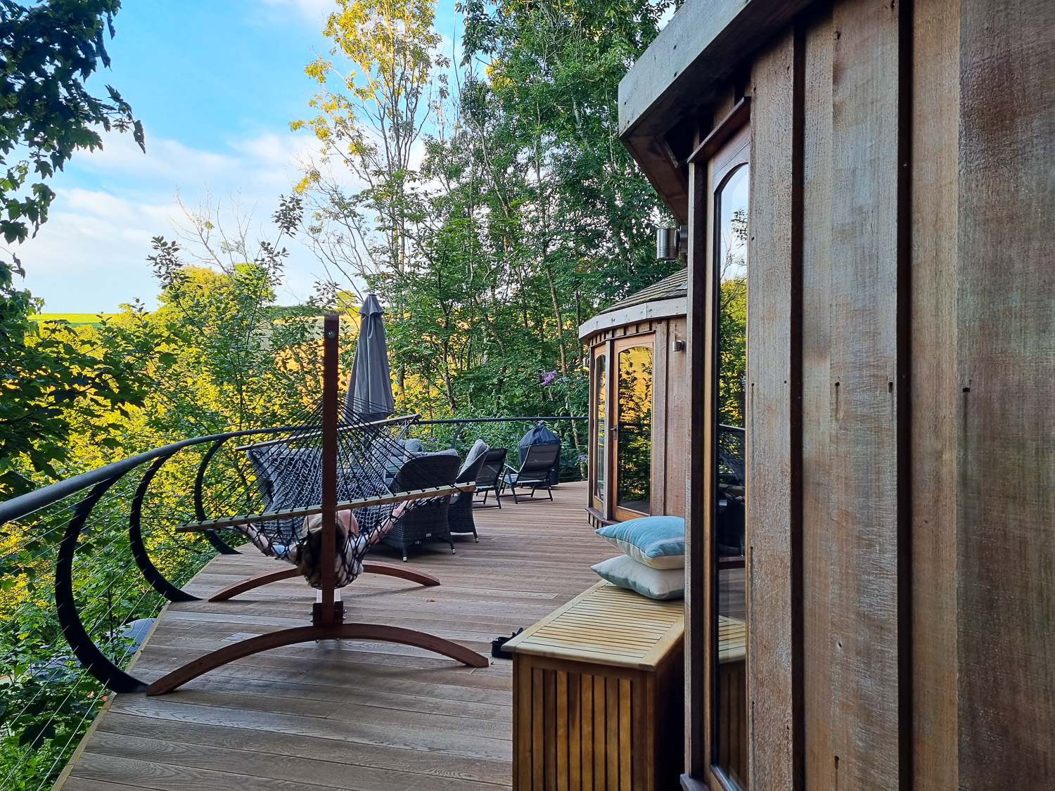 View of the hammock and wooden bench towards the rounded wooden exterior of Owl treehouse, with table, chairs and loungers on the deck beyond - my Dittisham Hideaway review after a stay at the Devon luxury glamping site