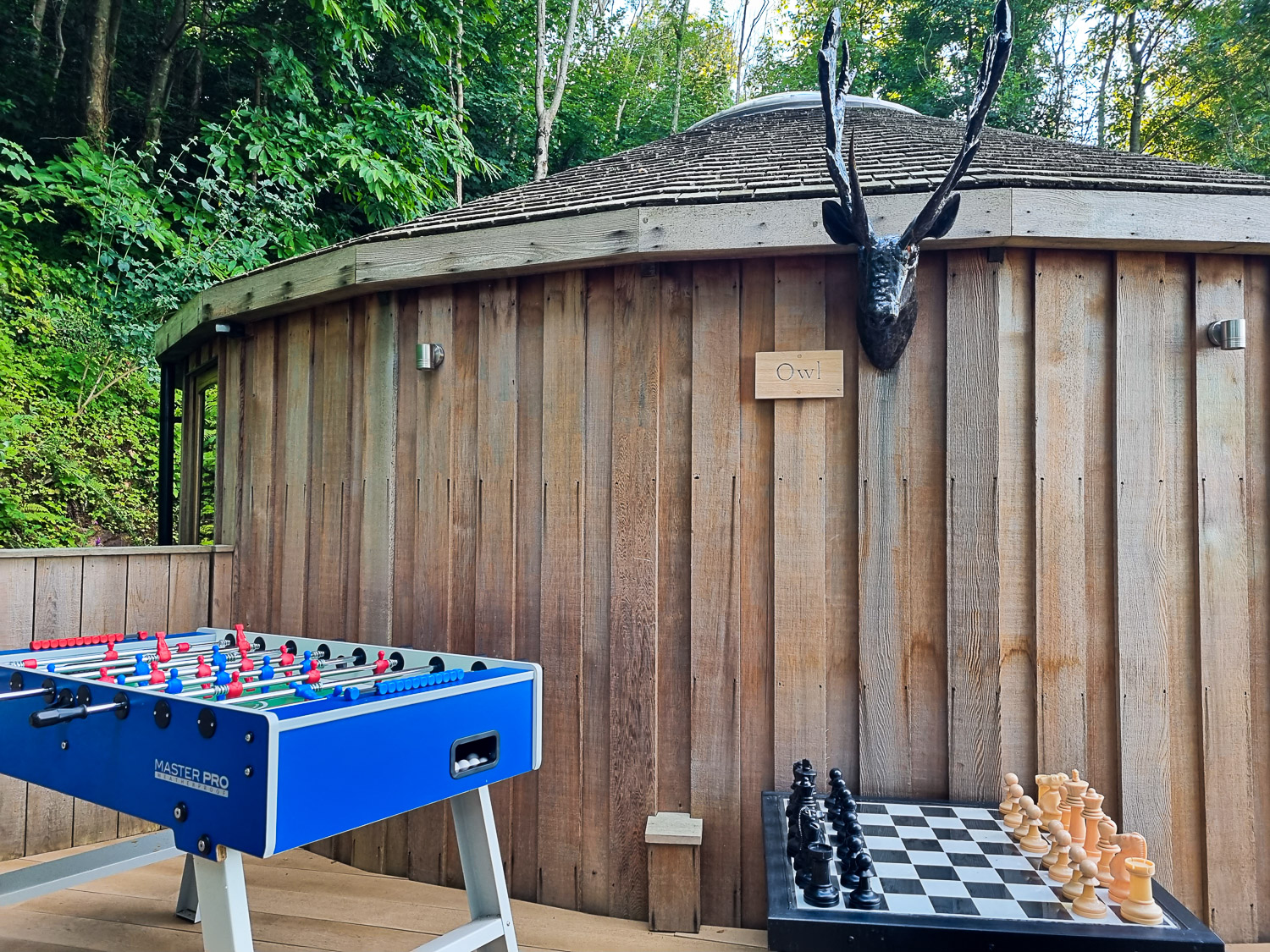 Side view of the wooden exterior of owl treehouse with a metal stag head above a table football and outdoor chess set - perfect for a stay at Dittisham Hideaway with kids
