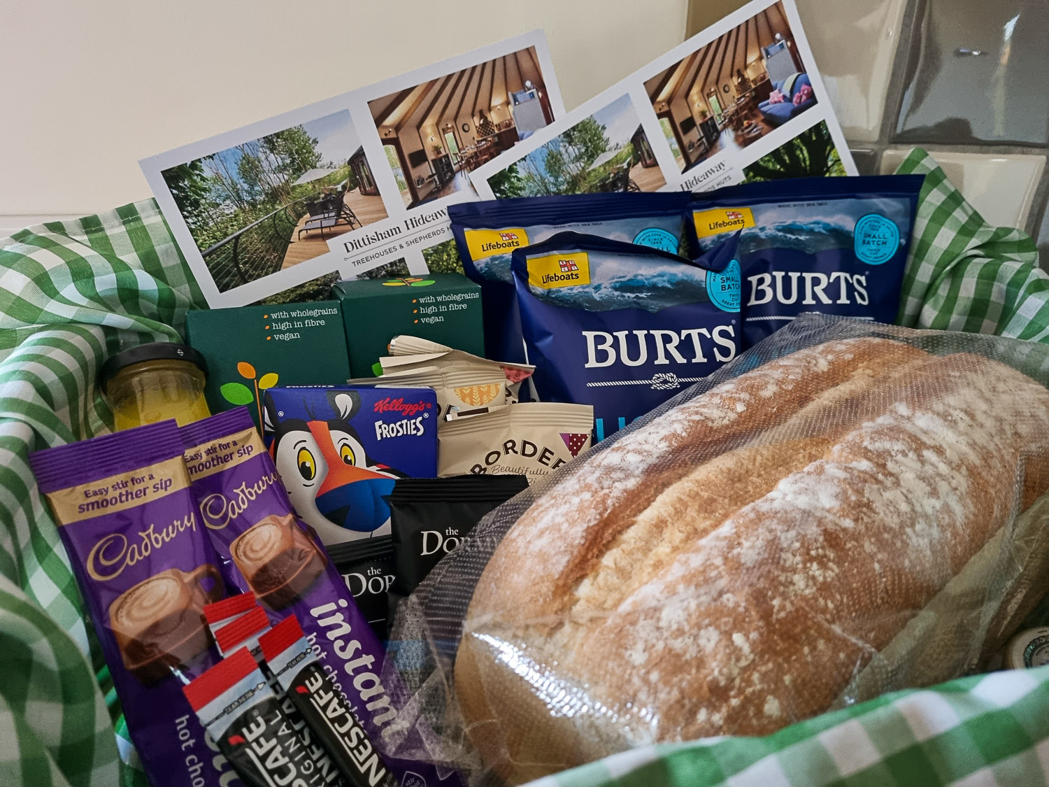 View of the hamper left for us at Owl Treehouse, including bread, biscuits, crisps and juice on green and white chequered cloth - my Dittisham Hideaway review