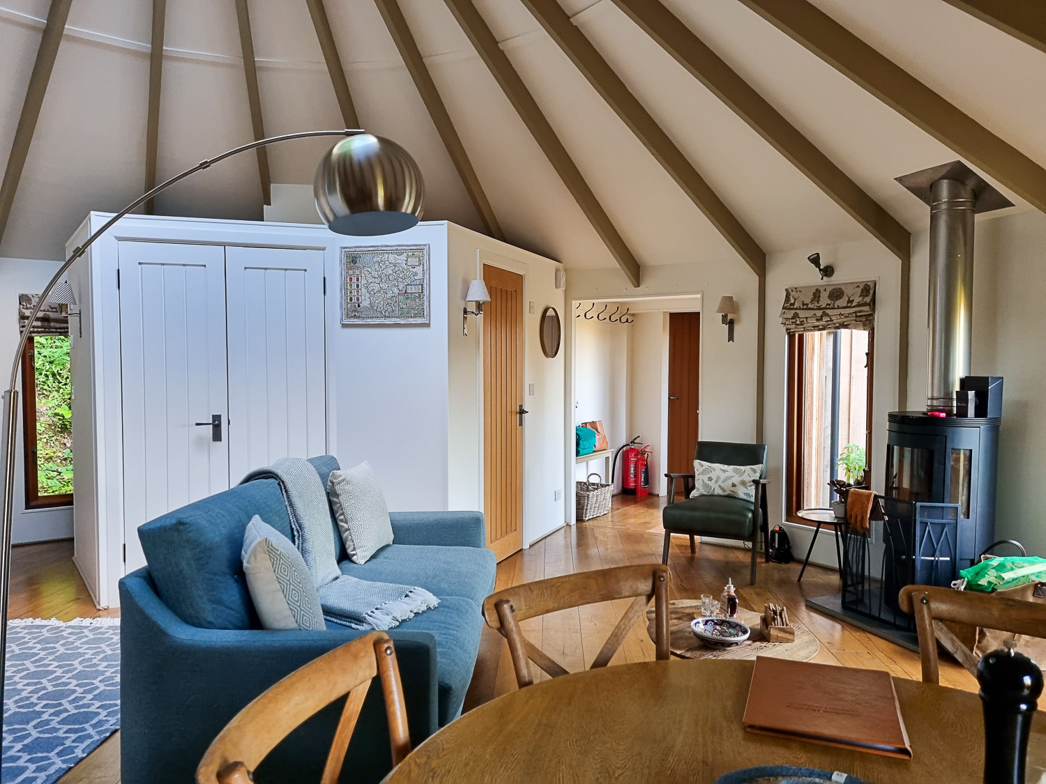 View across the wooden dining table to the blue armchair, wood-fired stove, chair and other rooms beyond at owl treehouse - my review of Dittisham Hideaway luxury glamping in Devon