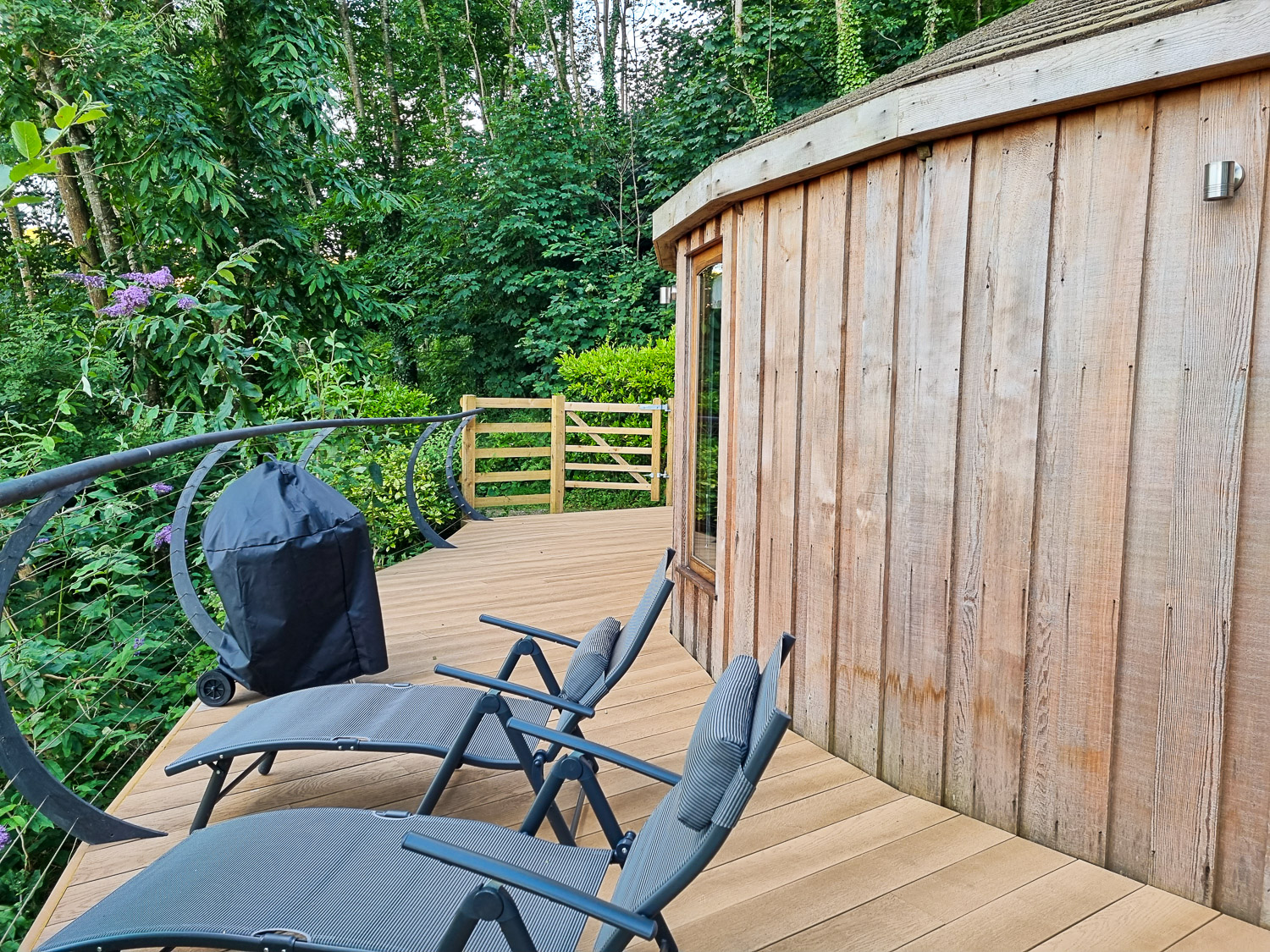View of the loungers and covered BBQ, as well as a side view of the rounded wooden exterior of Owl treehouse, taken from the deck - my review of Dittisham Hideaway luxury glamping in Devon