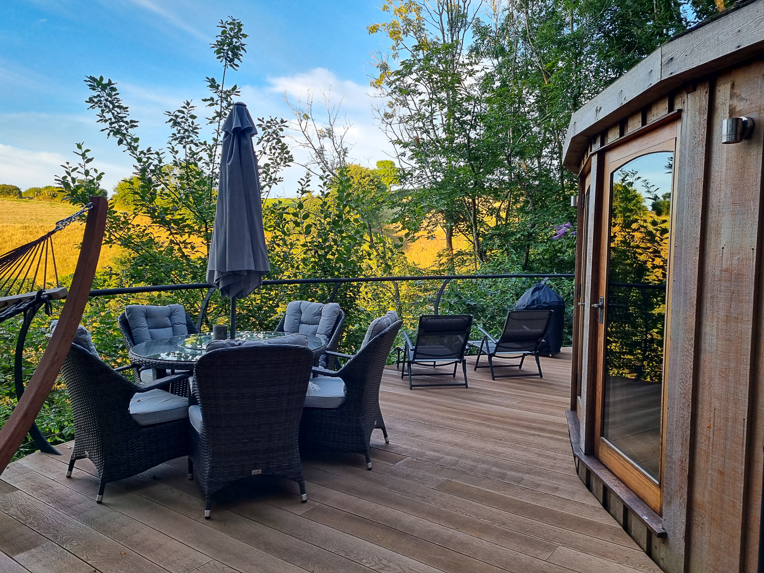 View of the rounded wooden exterior of Owl treehouse, with table, chairs and loungers on the deck to the trees and yellow fields beyond - my Dittisham Hideaway review after a stay at the Devon luxury glamping site