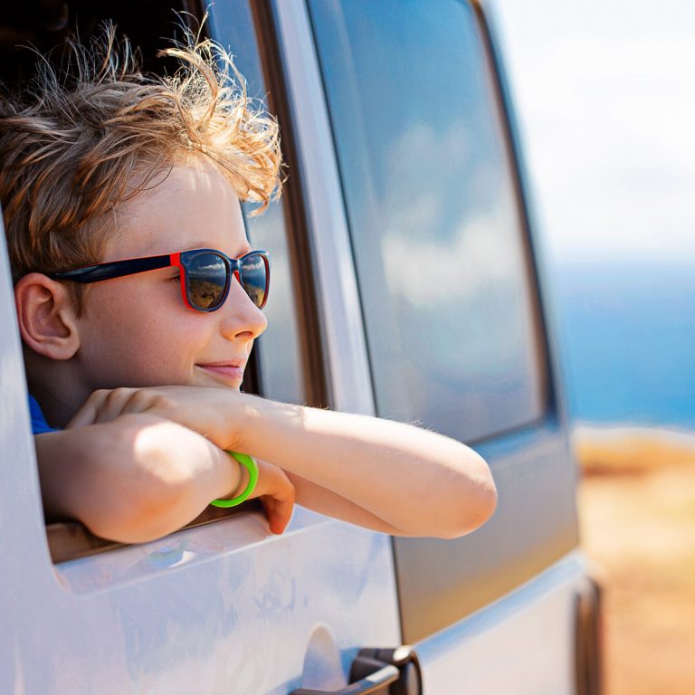 Boy with folded arms looking out of the rear window of a silver car with sea in the background - my tips if you're planning a road trip with kids