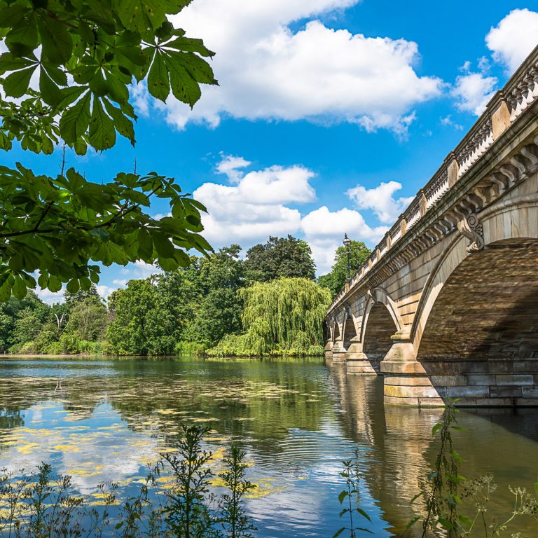 View across the water of the Serpentine in Hyde Park to the bridge with trees reflected in the water - where to cool down during a heatwave, including London attractions with air conditioning