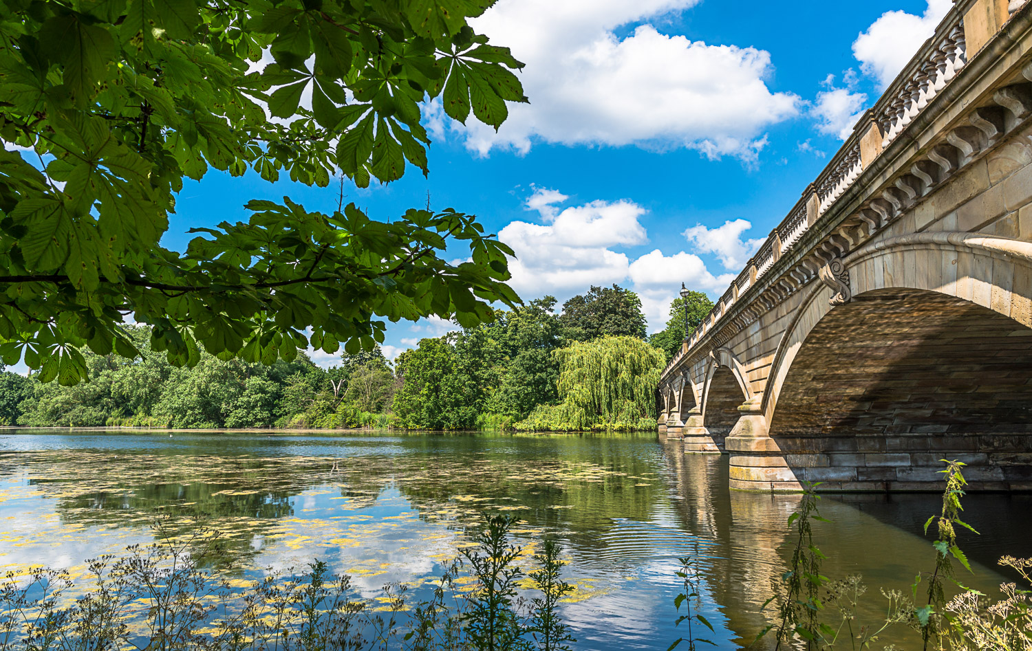 View across the water of the Serpentine in Hyde Park to the bridge with trees reflected in the water - where to cool down during a heatwave, including London attractions with air conditioning