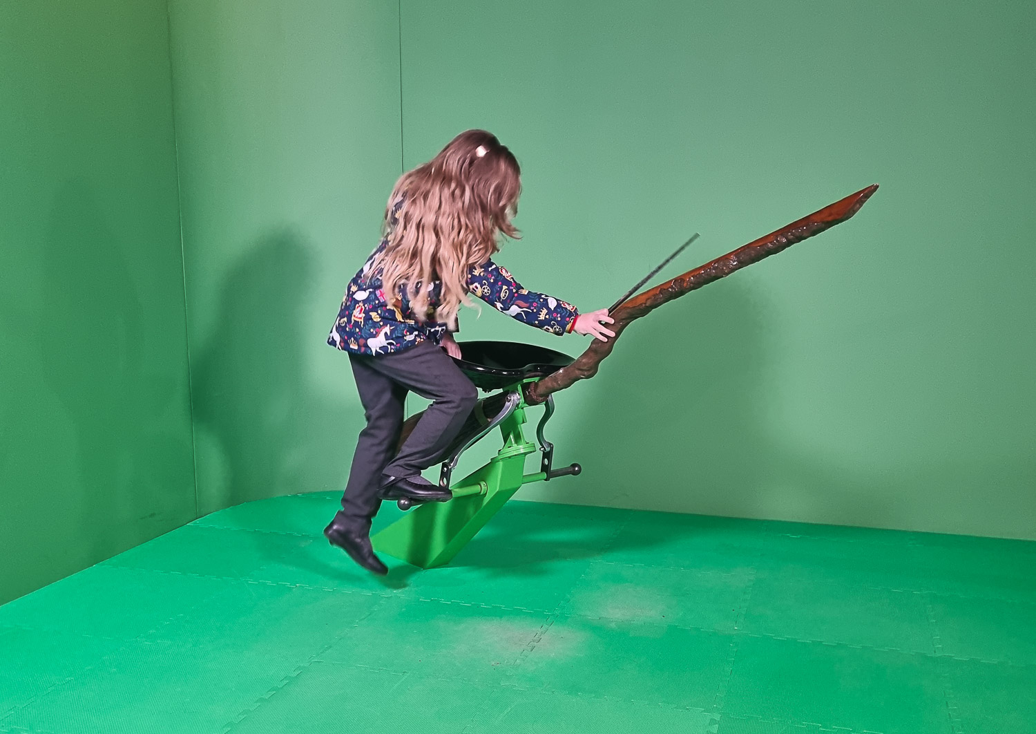 My daughter climbs onto a broomstick against a green screen while holding a prop wand - my review of London's Harry Potter Photo Exhibition with kids