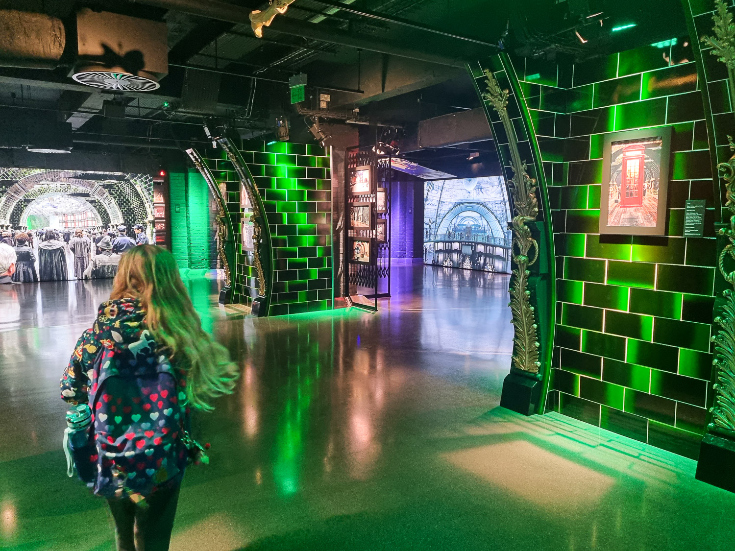 My daughter walks past displays in a green tiled section mimicking the Ministry of Magic during a visit to London's Harry Potter Photo Exhibition with kids