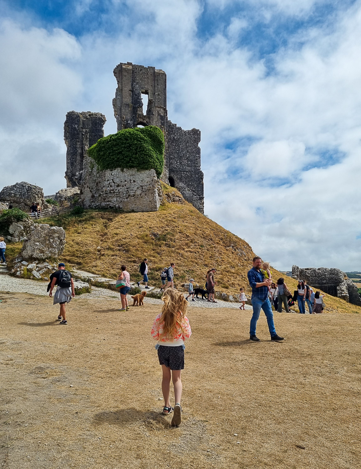 My daughter walks across the grass towards the hill where the highest ruins of Corfe Castle stand during a summer family holiday in Dorset