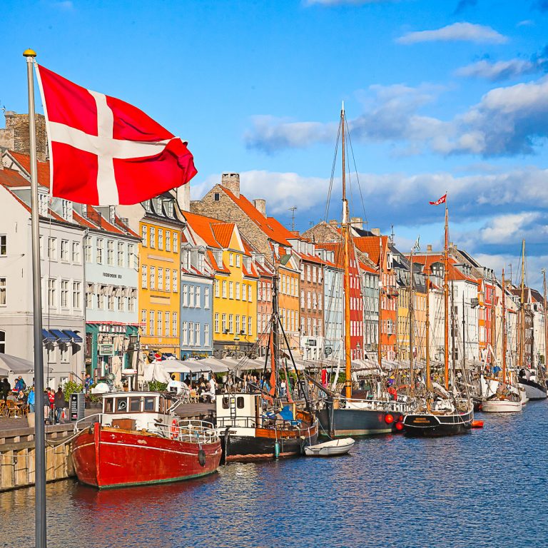 View of the Danish flag with boats and the colourful houses of Nyhavn in Copenhagen behind - my guide to Denmark with kids