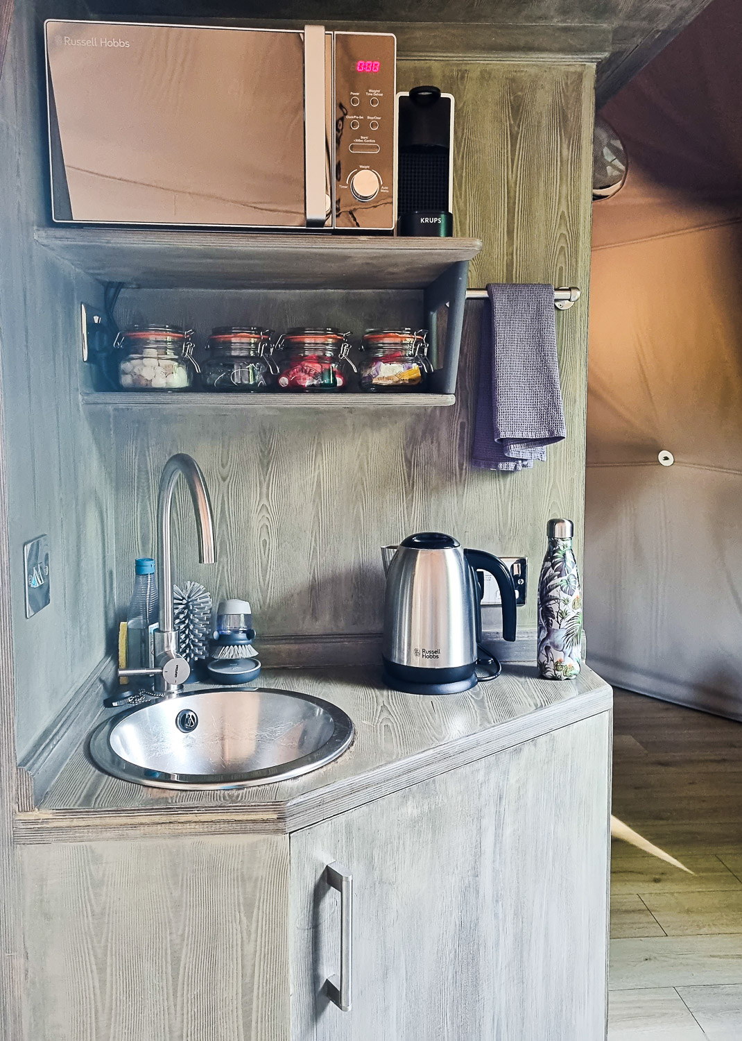 Small kitchenette area in Knole Luna Dome, with microwave, kettle, shelf of jars and sink above the cupboard - a luxury glamping break in Kent with kids