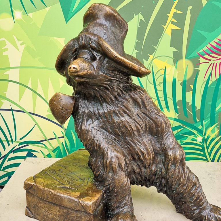 Bronze statue of Paddington Bear sitting on a suitcase against a leafy background at Paddington Station - one of several reproductions if you're trying to find Paddington Bear in London with kids