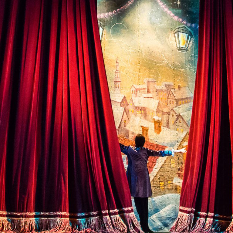 Male figure opens red theatre curtains to reveal a painted scene - the best Christmas theatre in London with kids, including pantomime, Christmas ballet, festive musicals, circus performances and more