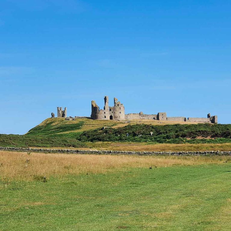 Ruins of Dunstanburgh Castle against a blue sky, seen across the fields from the clifftop path from Craster - visiting Dunstanburgh Castle Northumberland with kids