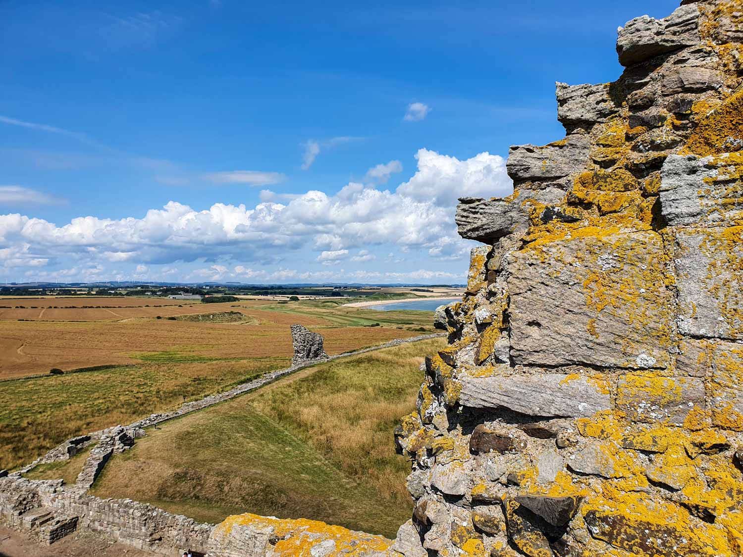 View around a crumbling grey stone wall covered with yellowing lichen to the stone wall and path down to Embleton Bay - visiting Dunstanburgh Castle with kids