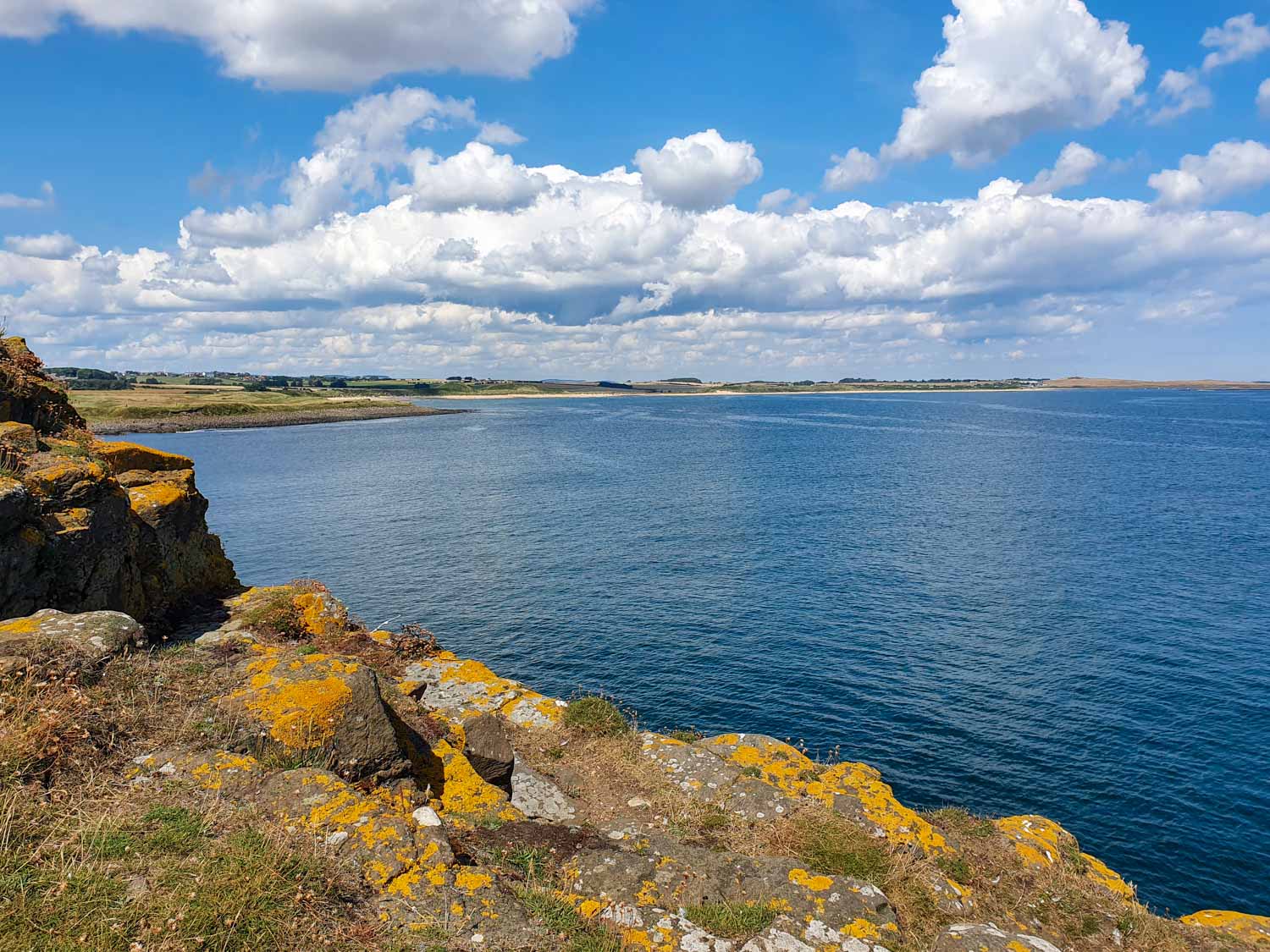 View over lichen covered stones to the deep blue waves of the sea and Embleton beach beyond visiting Dunstanburgh Castle Northumberland with kids